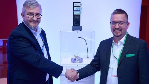 Two men in suits and glasses: Robert Pękal - the Director of PSNC and Thomas Monz - CEO and Founder of Alpine Quantum Technologies are shaking hands in front of a display case. Inside the illuminated case, a small, intricate metallic object rests on a circular platform. Behind the case, a tall, dark award or trophy with several logos including "PIAST-Q", "PSNC", and "AQT" is visible. The background features a softly lit blue and white backdrop. The overall setting suggests a formal event, possibly an award ceremony or a business interaction.