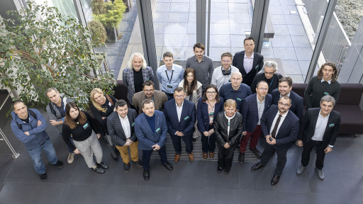 The image captures a group of approximately 25 adults posing for a photo from an elevated perspective. They are arranged in a somewhat staggered formation across a modern indoor space with large windows and natural light. Some individuals are standing while others are seated on a low bench. A leafy plant is visible on the left side of the frame, adding a touch of greenery to the setting. The overall atmosphere suggests a group gathering, possibly for a conference or a team photograph.
