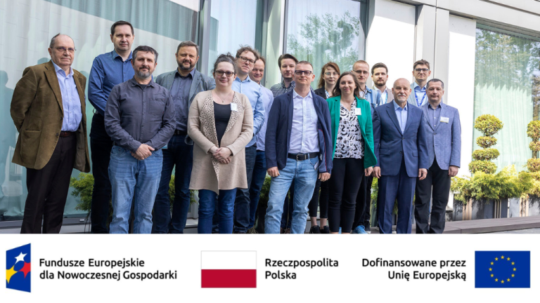 The picture shows a group of sixteen adults standing outdoors in front of the PSNC headquarters in Poznań. They appear to be posing for a photograph. Most of the individuals are looking towards the camera and some are smiling. The group is diverse in terms of clothing styles. Some individuals are wearing casual attire like sweaters and jeans, while others are dressed more formally in suits or blazers. At the bottom of the image, there are logos and text in Polish. From left to right, there is a logo with text that reads "Fundusze Europejskie dla Nowoczesnej Gospodarki" (European Funds for a Modern Economy). Next to it, there is the Polish flag and the text "Rzeczpospolita Polska" (the Republic of Poland). On the right side, there is text that says "Dofinansowane przez Unię Europejską" (Co-financed by the European Union) along with the European Union flag. These indicate that the project or event is likely funded by the European Union and related to Poland's modern economy. The background shows a well-lit exterior with some greenery visible. The overall impression is that this is a photograph taken at a project-related gathering.