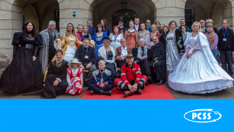This photograph captures a large group of people, many dressed in historical or elaborate costumes, gathered outdoors in front of a classical building. A red carpet is laid out in the foreground where some individuals are seated, while others stand behind them. The costumes vary, including formal gowns, military-style uniforms, and traditional attire, suggesting a historical reenactment, themed event, or performance. A blue banner with a white logo of PCSS is visible at the bottom of the image.