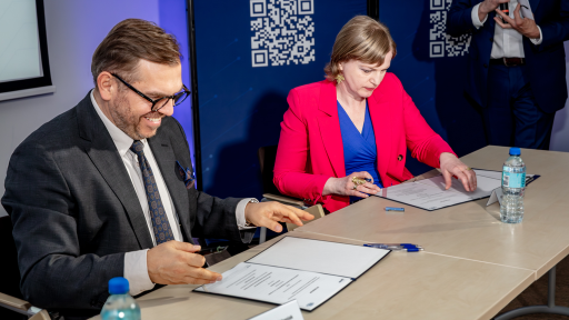 Two individuals, a man in a dark suit and glasses and a woman in a bright pink blazer and blue top, are seated at a light-colored table signing documents. Both appear focused on the papers in front of them, with the man smiling slightly. A water bottle and pens are also on the table. Behind them there is a blue wall with white QR codes and a partially visible third person in a dark suit.