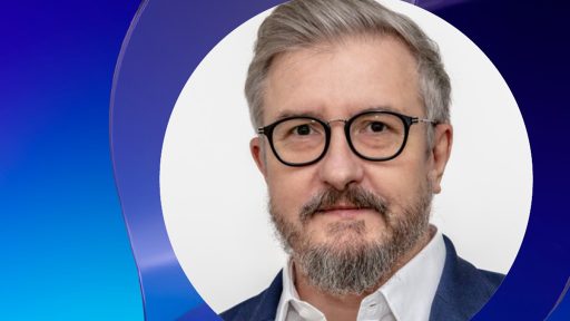 This is a portrait of a man with graying hair and a beard, wearing round black-rimmed glasses, a white collared shirt, and a blue blazer. His expression is neutral as he looks directly at the camera. The man is Robert Pękal – the Director of PSNC. The portrait is set against a backdrop that transitions from a lighter blue on the left to a darker blue on the right, with curved, layered shapes adding a dynamic element.