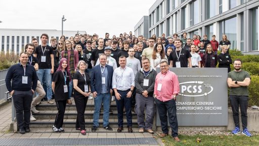 A large group of people, mostly young adults, are gathered outside a building with a sign that includes "PCSS" and "Poznańskie Centrum Superkomputerowo-Sieciowe." The group is diverse and casually dressed, with some individuals standing on steps leading up to the building and others on the ground. Several people in the front row appear to be slightly older and could be organizers or staff.