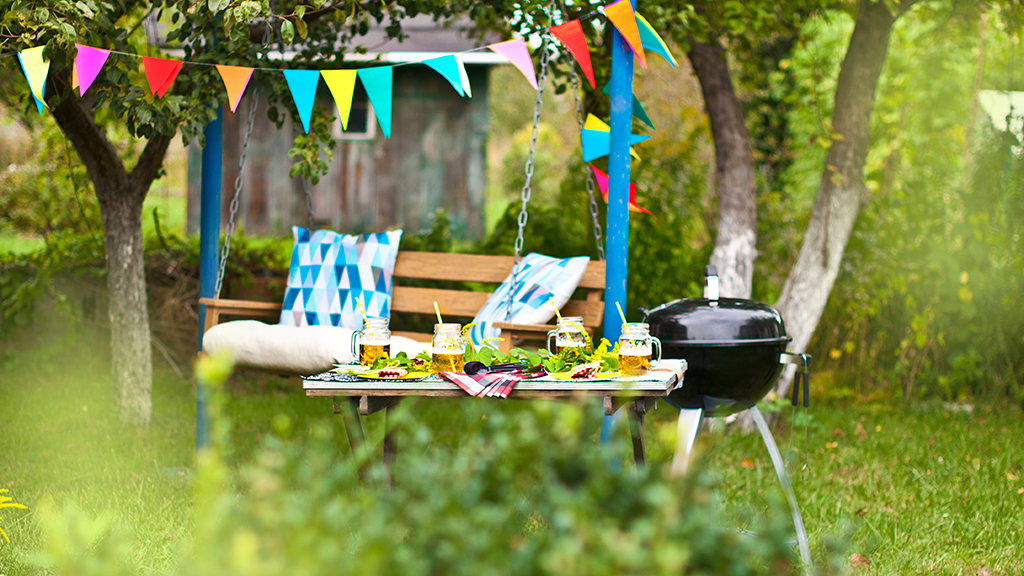 PPAM 2024 The image shows a "Majówka" scene, featuring a decorated swing with colorful flags hanging between trees. A table is set with food and drinks and a grill stands nearby, suggesting an outdoor celebration. The setting appears to be a garden or backyard, creating a festive atmosphere.