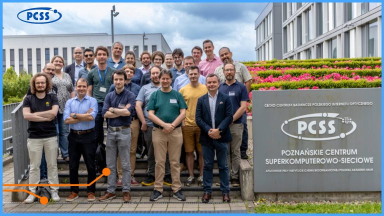 The image shows a large group of people, predominantly men, posing for a photo outdoors on a set of steps in front of a modern building. Most individuals are smiling and appear relaxed, dressed in a mix of smart casual and casual attire. To the right side of the image, there is a gray sign displaying the "PCSS" logo and text that reads "POZNAŃSKIE CENTRUM SUPERKOMPUTEROWO-SIECIOWE" (Poznań Supercomputing and Networking Center) and "CENTRUM BADAWCZE POLSKIEGO INTERNETU OPTYCZNEGO" (Polish Optical Internet Research Centre). The background features green foliage, blooming pink flowers on a sloped garden bed, and parts of other buildings under a cloudy sky.