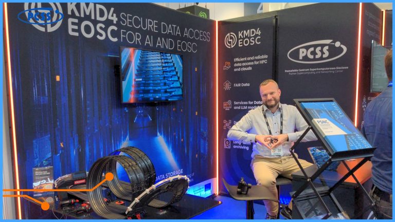 The photo depicts a conference booth at the ISC25 conference in Hamburg. In the center of attention is a bearded man, dressed in a light-colored shirt and beige trousers, sitting on a chair and looking directly at the camera. In the background, banners with the text "KMD4 SECURE DATA ACCESS EOSC FOR AI AND EOSC" and "PCSS Poznańskie Centrum Superkomputerowo-Sieciowe" are visible, suggesting the booth is related to data access in the context of artificial intelligence and EOSC. On the table in front of the man, there is a small racetrack with toy cars, and monitors display graphics related to data and supercomputers.