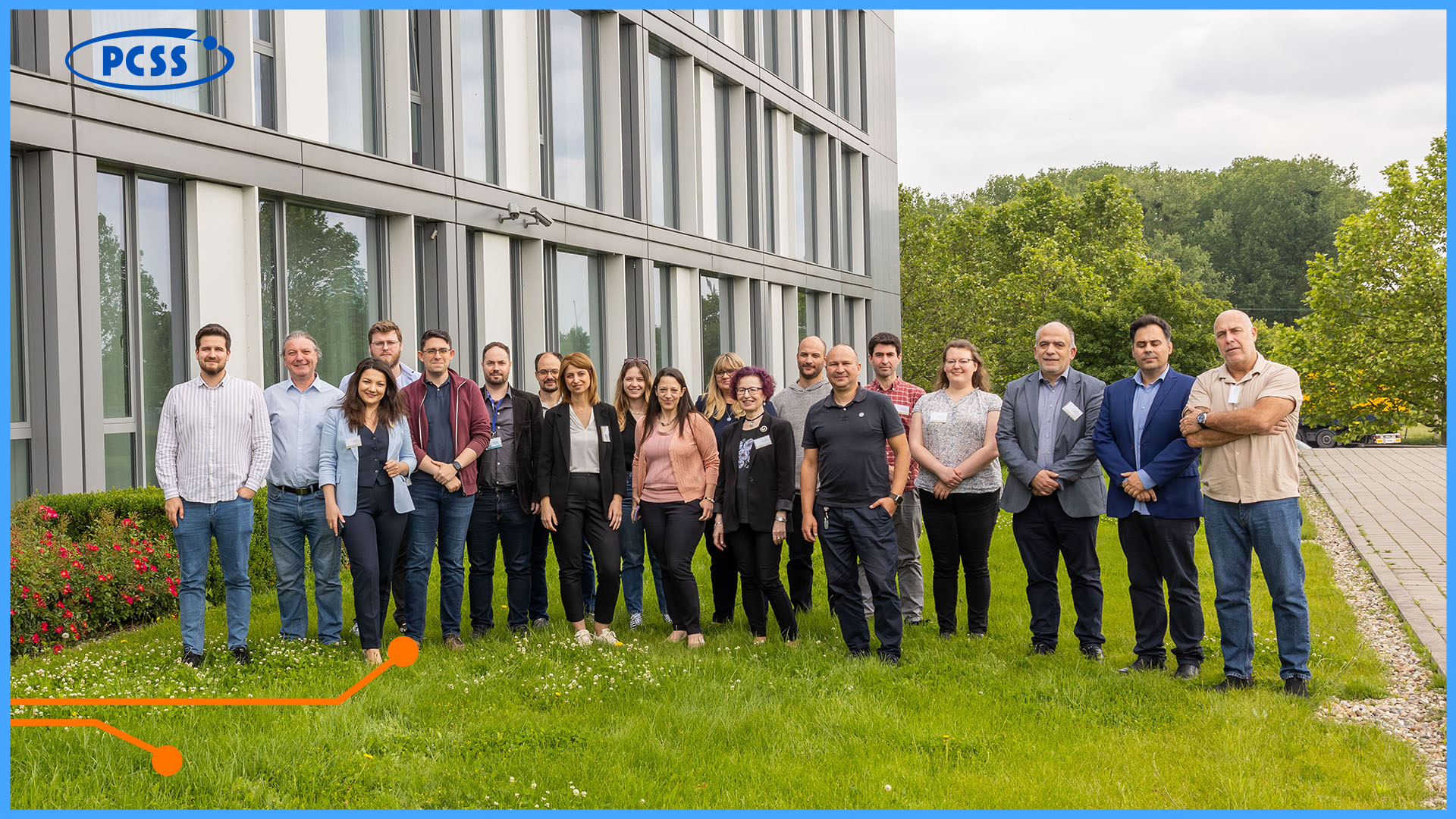 The image displays a group of approximately 20 adults, standing on a lush green lawn in front of a modern building with large windows. The individuals are casually dressed, with most facing the camera and some engaged in conversations, suggesting a relaxed professional gathering. In the top left corner, a logo of PCSS is visible, indicating the affiliation of the location.