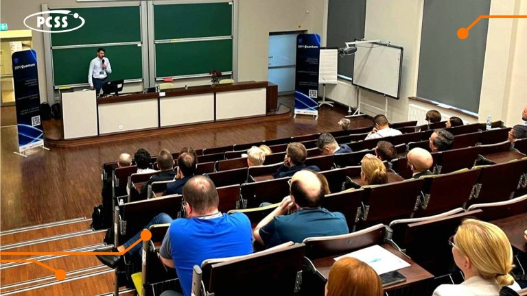 A high-angle, wide shot shows a conference speaker standing at a podium in a lecture hall, facing an audience. The speaker, positioned at the front left, addresses attendees seated in rows of wooden desks and chairs. Two large green chalkboards are visible behind the speaker, and a projection screen is set up on the right side of the stage.